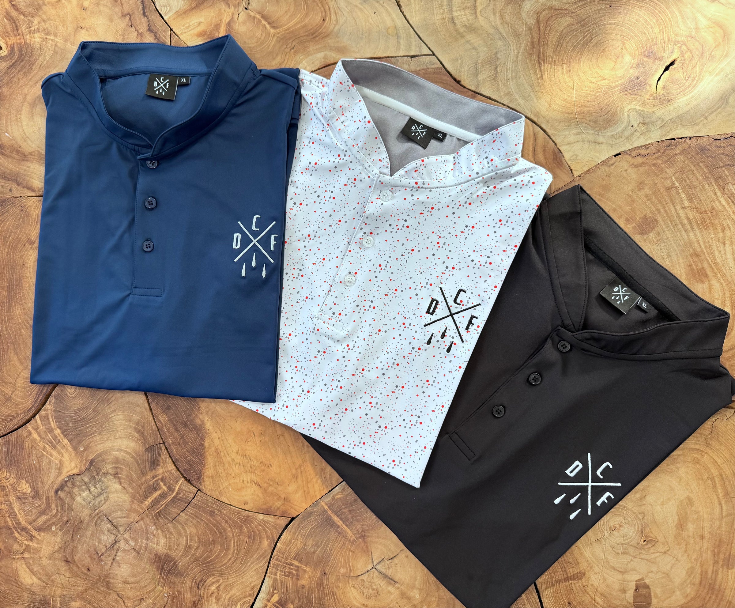 Three polo shirts in blue, white with red speckles, and black on a wooden surface.